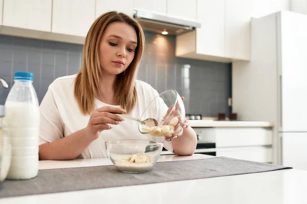 Το πρωινό είναι μεγάλη υπόθεση. Νεαρή γυναίκα προσθέτει μπανάνα σε ένα μπολ με πλιγούρι βρώμης ενώ ετοιμάζει πρωινό στην κουζίνα — Φωτογραφία Αρχείου
