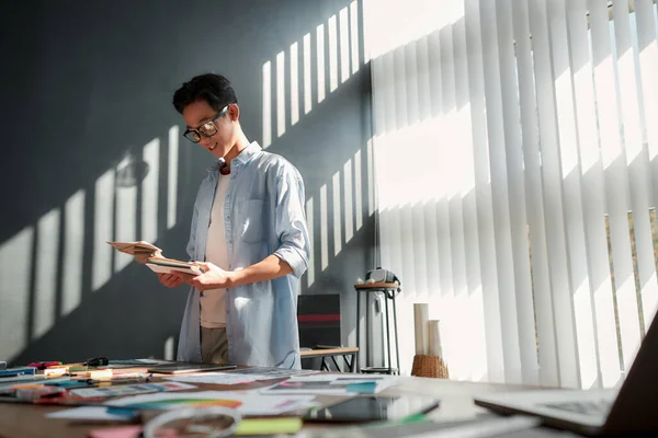 Working on a new project. Smiling asian man in eyeglasses looking at color palette while standing near the office table with a lot of creative stuff on it — Stock Photo, Image