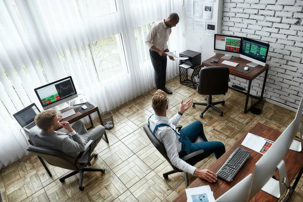 A world of business. High-angle view of three traders having chat while sitting by desks in front of computer monitors. Diverse employees working in the office. — Stock Photo, Image