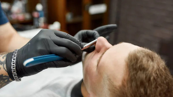 Небезпечна бритва. Чоловічий перукар в чорних рукавичках з татуйованою рукою гоління клієнта з прямою бритвою. Традиційне гоління. Веб банер — стокове фото