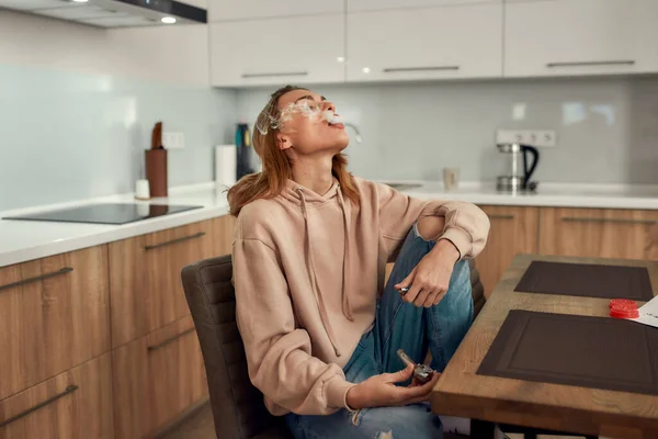 No Bad Vibes. Young caucasian woman exhaling the smoke while smoking marijuana from a metal pipe, sitting in the kitchen. Red weed grinder on the table — Stock Photo, Image