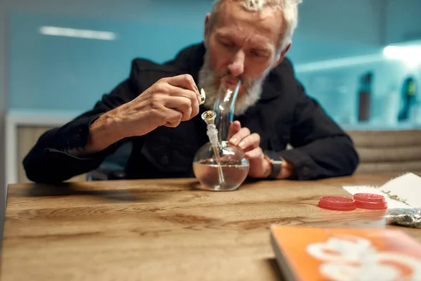 À procura de relaxamento. Homem sênior acendendo cannabis na tigela de tubo de água de vidro ou bong na cozinha. Ferramentas de marijuana na mesa. Legalização e dependência do conceito de drogas leves — Fotografia de Stock