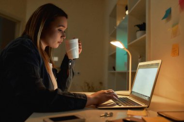 Drinking coffee for some energy. Side view of young woman working from home in the evening or night. She is using laptop, typing on keyboard clipart