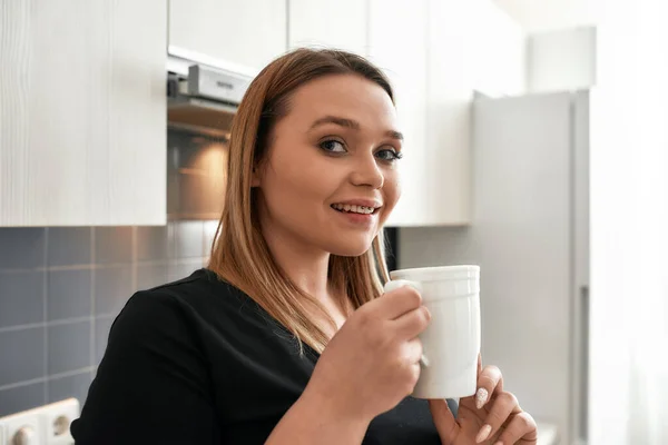 Kaffee macht alles möglich. Nahaufnahme einer jungen kurvigen Frau, die in die Kamera schaut, während sie Tee oder Kaffee in der Küche trinkt — Stockfoto