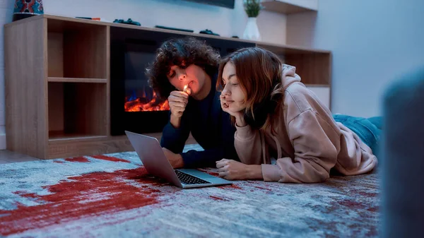 Disfrutando de la marihuana. Joven pareja de adolescentes fumando marihuana y mirando a la computadora portátil, tendida en el suelo en casa. Tipo encendiendo cigarrillo de cannabis — Foto de Stock