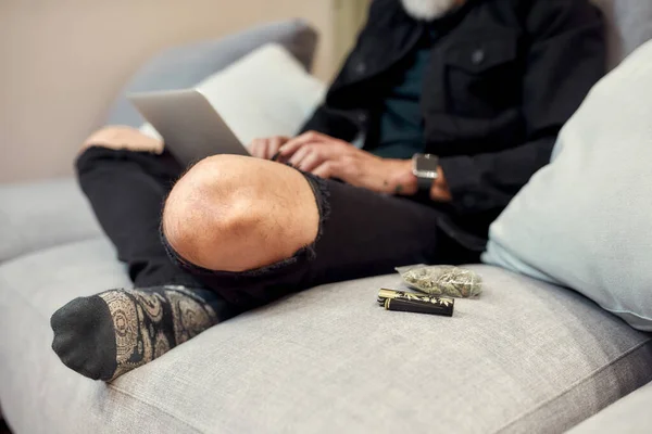 Para maior produtividade. Feche os botões no saco de plástico e leve deitado no sofá. Um homem a usar um portátil ao fundo. Cannabis e conceito de legalização de ervas daninhas — Fotografia de Stock