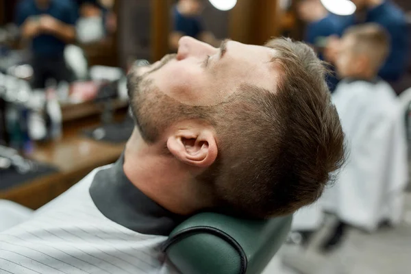 Bartkorrektur. Seitenansicht eines gutaussehenden bärtigen Mannes, der im Friseurstuhl liegt und sich beim Friseur rasiert. Rasierkonzept — Stockfoto