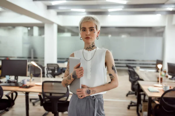 Smart business lady. Portrait of young, confident and attractive tattooed business woman holding her laptop and looking at camera while standing in the modern coworking space