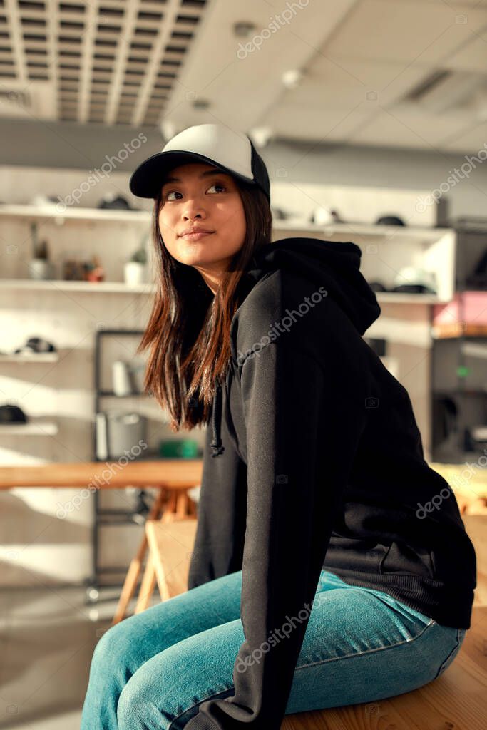 Cheerful female worker looking aside while posing in custom apparel, baseball cap and hoodie. Young woman working at custom T-shirt, clothing printing company. Vertical shot