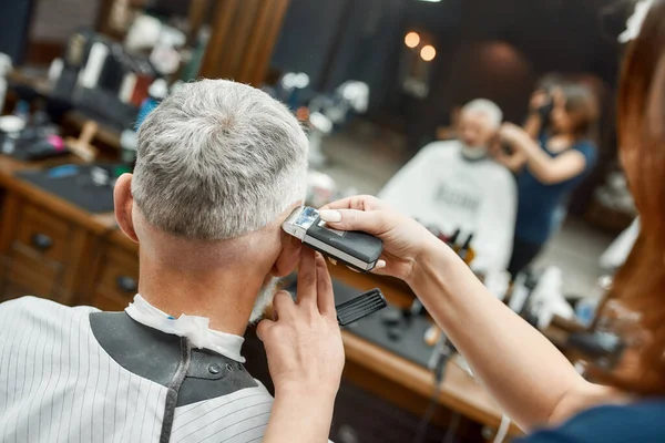 Vista posteriore dell'uomo maturo seduto sulla sedia del barbiere mentre il barbiere femminile lavora con il tagliacapelli, rendendo il taglio di capelli. Barbiere. Focus selettivo — Foto Stock