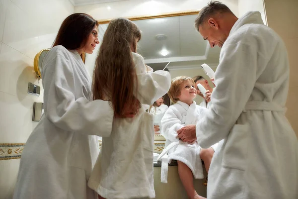 Todo el mundo necesita cuidado. Los padres caucásicos y dos niños en batas blancas que tienen belleza, rutina de la mañana en el baño del hotel, de pie cerca del espejo. Familia, viajes, resort, concepto de vacaciones — Foto de Stock