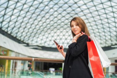 Bulanık alışveriş merkezindeki telefon ve alışveriş torbalarıyla güzel, koyu saçlı kız.
