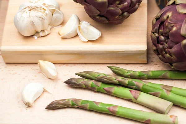 Artichokes, asparagus stems and garlic on a wooden kitchen board — Stock Photo, Image