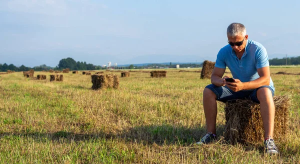 Un hombre sentado en un campo con un teléfono en un pajar . — Foto de Stock