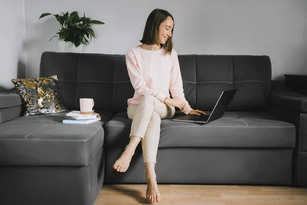 Молодая женщина сидит на диване и использует ноутбук . Стоковое Изображение