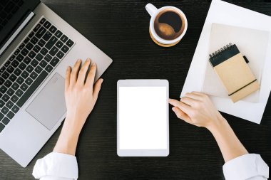 Üst görüntüde beyaz tablet elinde kadın eli resmi var. Beyaz masaüstü ekranı dizüstü bilgisayarı ve kahve fincanları evdeki ya da ofisteki ahşap siyah masada..