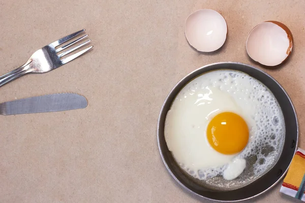 Rzemieślnicze tło z jajkiem smażonym na patelni, widelec i nife, skorupa jajka. — Zdjęcie stockowe