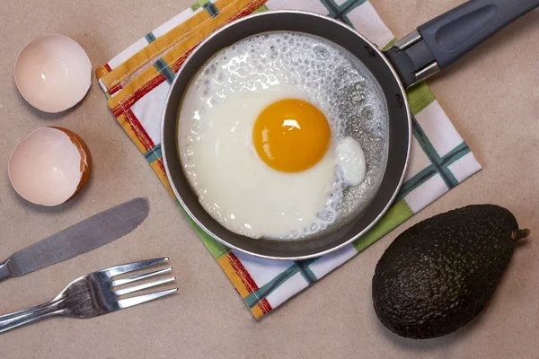 Jajko smażone na patelni, widelec i nóż, awokado, skorupki jaj, ręcznik do naczyń. — Zdjęcie stockowe