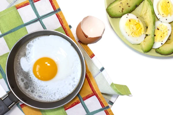 Smażone jajko na patelni, zielony talerz z gotowanym jajkiem i awokado, ręcznik kuchenny na białym stole — Zdjęcie stockowe
