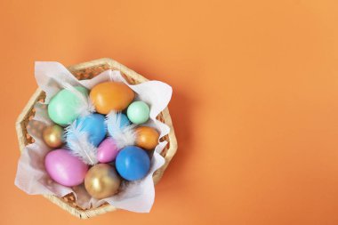 Turuncu arka plandaki hasır sepette altın, pembe, mavi, yeşil, turuncu yumurtalar. Uzayı kopyala Metin ve tasarım için yer. Mutlu Paskalyalar. Üst Manzara. Düz Düzen.