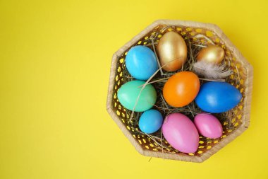 Sarı arka plandaki hasır sepette altın, pembe, mavi, yeşil, turuncu yumurtalar. Uzayı kopyala Metin ve tasarım için yer. Mutlu Paskalyalar. Üst Manzara. Düz Düzen.
