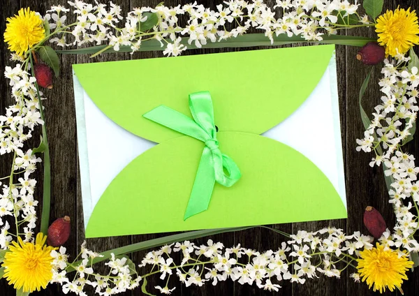 Green gift envelope with green ribbon in a frame of flowers of bird cherry, dandelions and berries of wild rose on wooden background. Flat lay.