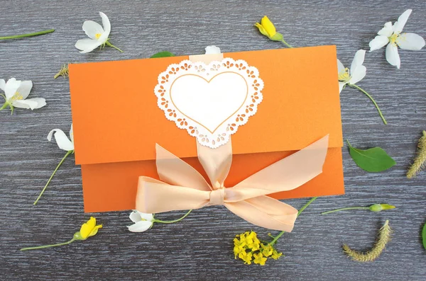 Orange gift envelope tied with a pink ribbon on wooden background with flowers.