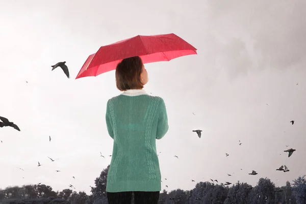 A young girl in a green sweater with a red umbrella stands in the rain and looks at the gloomy sky with birds. The concept of bad weather. Gothic rainy mood. rear view.