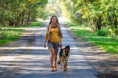 Mutlu bir kız ormanda bir Alman çoban köpeğiyle yolda yürüyor ve gülümsüyor. Genç bir köpek yavrusu metresini koruyan sadık bir dosttur. Ağaçların sarı-yeşil sonbahar arka planında.