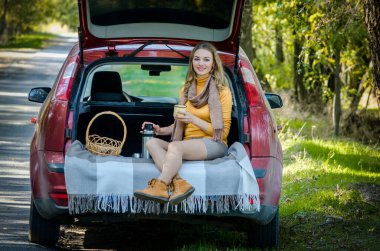 Güzel sarışın bir kız kırmızı bir arabanın bagajında oturuyor, gülümsüyor.,