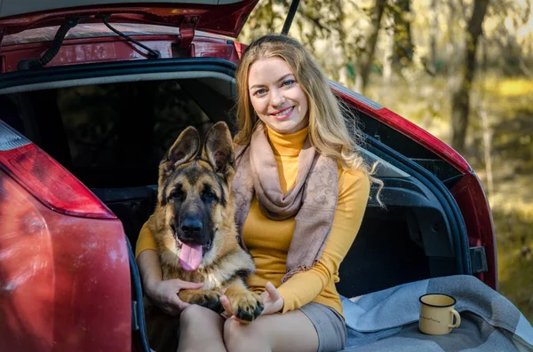 En glad vit flicka ligger i bakluckan på en bil med en tysk herde hund ras och leenden, en glad lekfull tonåring valp tittar på kameran. Vänskap mellan människa och djur, resor, camping. Kopiera utrymme — Stockfoto