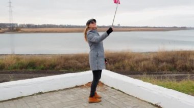 Kız seyahat blogcusu telefonda canlı yayında, yerel manzarayı gösteriyor. Selfie çubuğuyla video kaydediyor. Açık hava ceketli bir kadın gülümsüyor. Teknoloji ve yaşam tarzı kavramı.