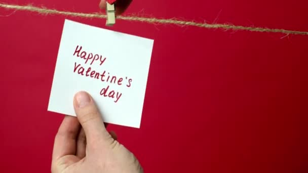 Närbild av en anteckning på ett rep med text glad Alla hjärtans dag. Flickornas händer hänger ett vitt papper på en röd bakgrund på en klädnypa. Begreppet kärleksförklaring, romantiska relationer — Stockvideo