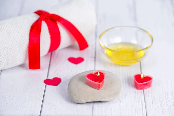 Koncept lázní na Valentýna. Červené svíčky ve tvaru srdce, kameny, masážní olej a bílý ručník na dřevěném pozadí. Relaxační a wellness péče. Koupací procedura. — Stock fotografie