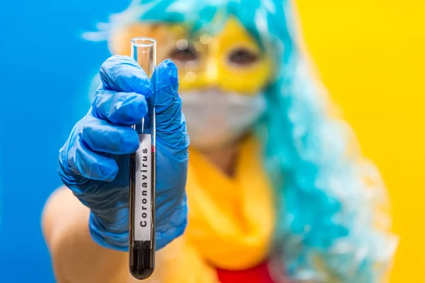 Concept de protection contre le coronavirus au carnaval. Une femme brillante avec une perruque bleue, un masque jaune et des gants médicaux tient un tube à essai avec un test sanguin pour la couronne du virus. Gros plan . — Photo