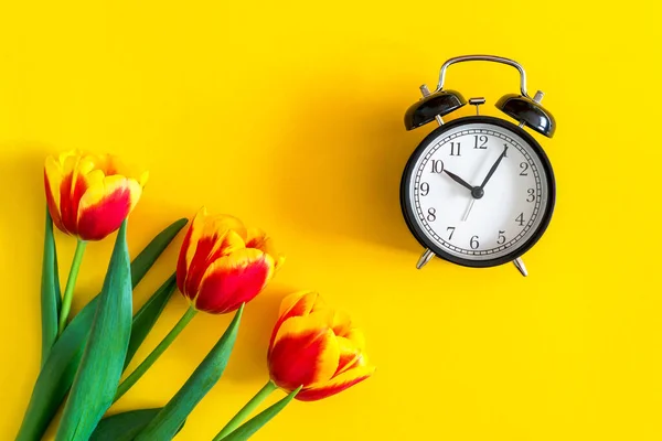 Concept time of spring, gifts for women and flowers. beautiful red and yellow fresh tulips and alarm clock on a bright yellow background. flat lay, top view. minimalistic greeting cards, spring mood.