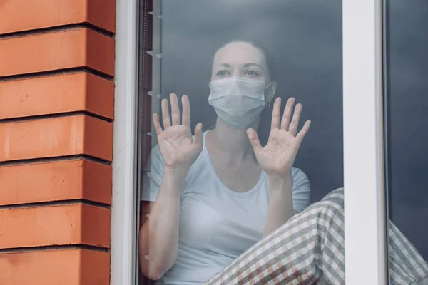 Quarentena em casa. Uma mulher caucasiana está sentada à janela com uma máscara médica, olhando para fora, sua mão em vidro, quer sair. proteção contra a infecção pelo coronavírus, pandemias, epidemias de surtos . — Fotografia de Stock