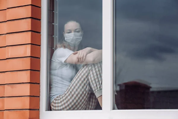 Quarentena em casa. Mulher branca sentada à janela com uma máscara médica, olhando para fora, quer sair. protecção contra a infecção pelo coronavírus, pandemias, surtos de doenças e epidemias. Espaço de cópia . — Fotografia de Stock