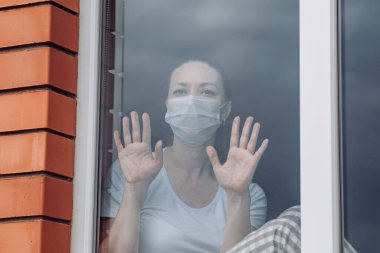 Ev karantinası. Kafkasyalı bir kadın pencerenin önünde tıbbi bir maskeyle oturuyor, dışarı bakıyor, eli camın üzerinde, dışarı çıkmak istiyor. Koronavirüs enfeksiyonuna karşı koruma, salgınlar, salgınlar.
