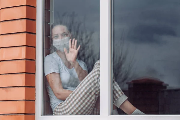 Quarentena em casa. Uma mulher caucasiana está sentada à janela com uma máscara médica, olhando para fora, sua mão em vidro, quer sair. proteção contra infecção por coronavírus, pandemias. Espaço de cópia . — Fotografia de Stock