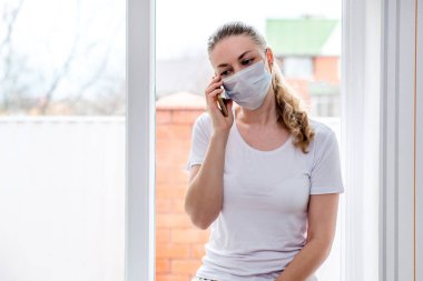 Ev karantinası. Sağlık koruma maskesi takmış beyaz bir kadın, telefon eder, ambulans çağırır. Kötü sağlık, yüksek sıcaklık, mevsimsel grip ve grip salgını. Ev tedavisi.