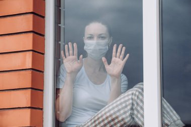 Ev karantinası. Kafkasyalı bir kadın pencerenin önünde tıbbi bir maskeyle oturuyor, dışarı bakıyor, eli camın üzerinde, dışarı çıkmak istiyor. Koronavirüs enfeksiyonuna karşı koruma, salgınlar, salgınlar
