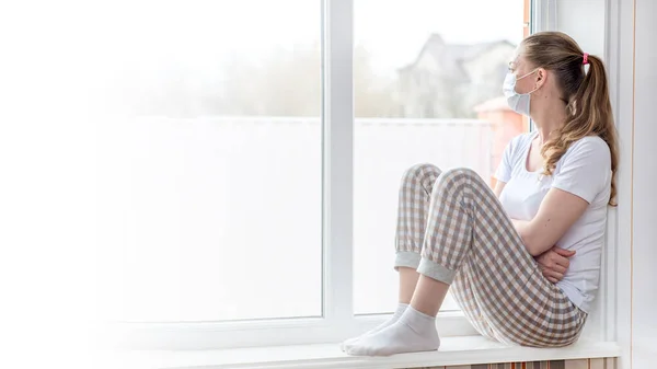 Home quarantine banner. A Caucasian woman on sick leave is sitting at the window in a medical protective mask, looking sadly at the street. Poor health, a seasonal epidemic of the flu virus and colds