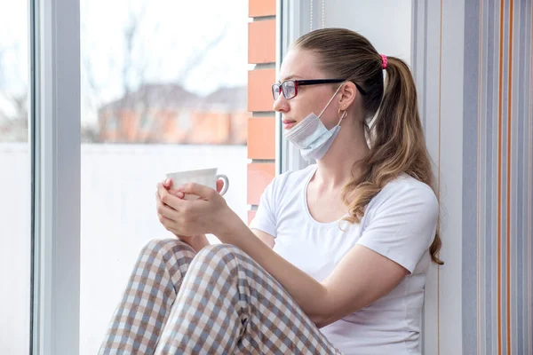 Home quarantine. Caucasian woman on sick leave is sitting at window in a medical protective mask, drinking medicinal herbal tea or water. Poor health, a seasonal epidemic of flu virus and colds