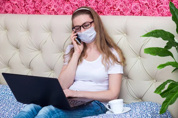 Remote work in home quarantine. A beautiful Caucasian girl is working on a laptop in a protective medical mask, talking on the phone, ordering food and medicine. Student on e-learning