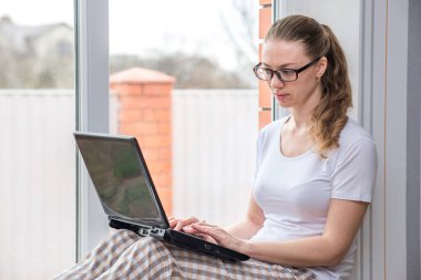 Ev karantinası. Uzaktan kumandalı iş. Koronavirüsten koruma. Penceresinde bilgisayar ve bir fincan kahve olan beyaz bir kadın. E-öğrenme ve arkadaşlık ve akrabalarla çevrimiçi iletişim