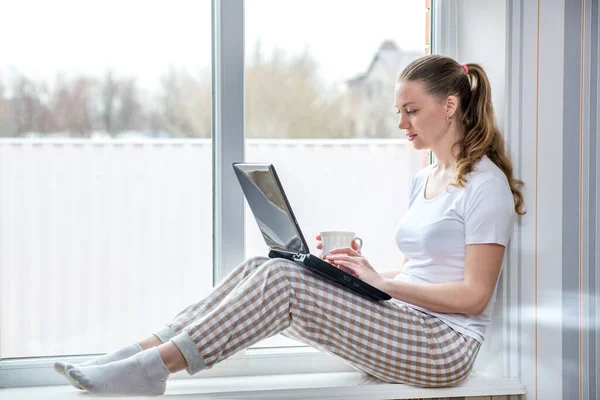 Home quarantine. Remote work at home. Protection from coronavirus. Caucasian woman at window with a computer and a Cup of coffee. E-learning and communicating online with friends and relatives