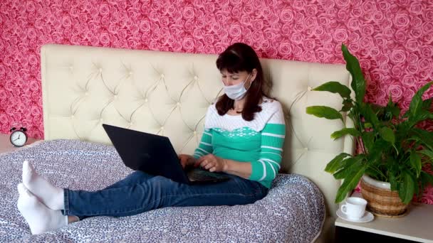 Remote work from home. An adult Caucasian woman is working on a laptop in her bedroom on a bed in a protective medical mask. The concept of business and e-education. Self-isolation in quarantine. — Stock Video