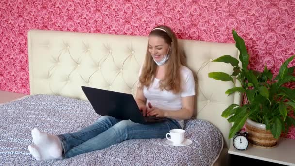 Fjärrarbete hemma. Vacker vit flicka arbetar på en bärbar dator i sitt sovrum på en säng i skyddande medicinsk mask och leende. Begreppet företag och e-utbildning. Självisolering i karantän. — Stockvideo