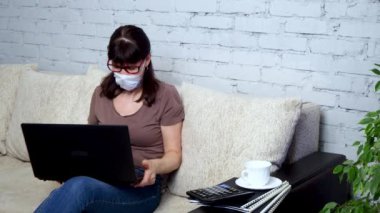 Evden uzak bir iş. Yetişkin bir beyaz kadın koruyucu bir tıbbi maskeyle bir kanepede dizüstü bilgisayar üzerinde çalışıyor. İş ve e-eğitim kavramı. Karantinada kendini izole etme.